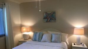 A neatly made bed with white bedding and blue pillows, two bedside lamps, a starfish artwork on the wall, and a window with curtains.