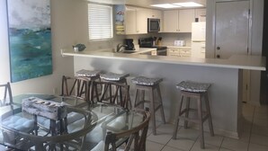 A compact kitchen with a central island, bar stools, a dining table, and a large abstract painting on the wall.