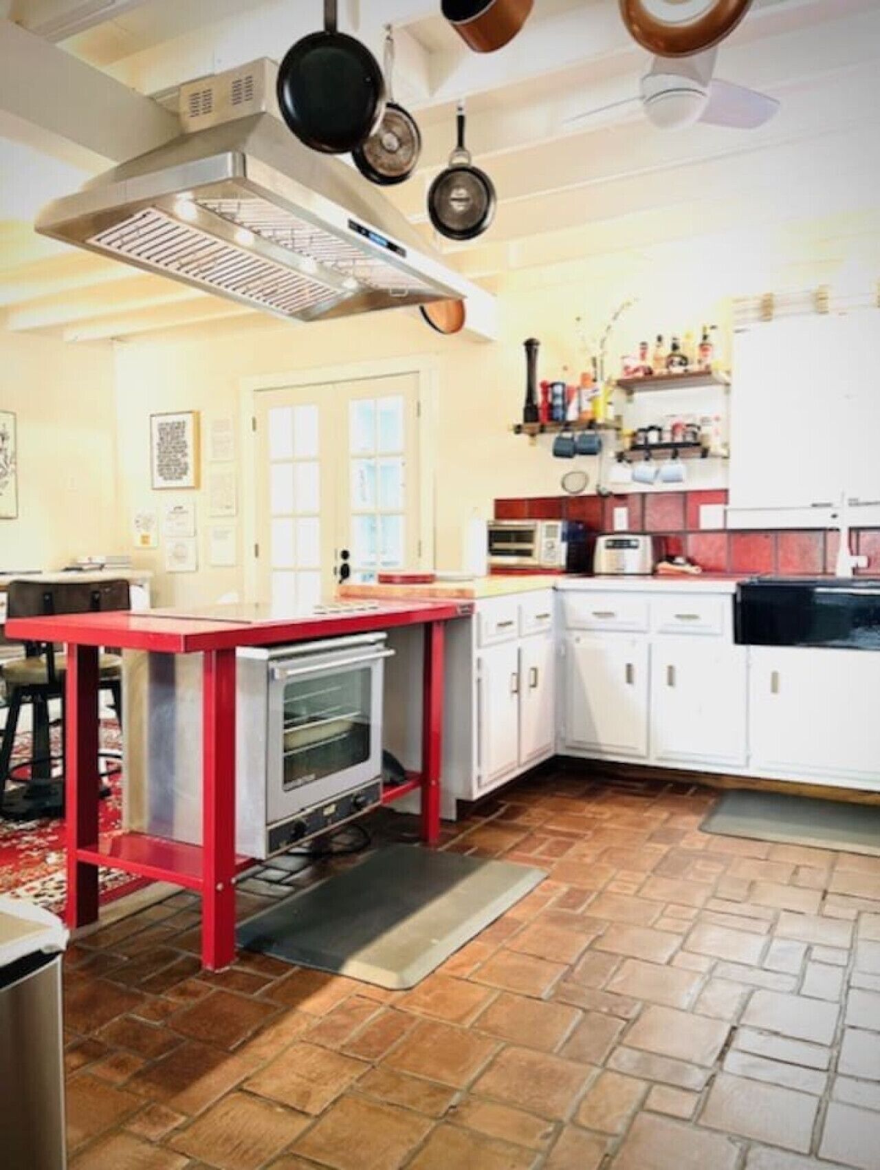 Une cuisine avec des meubles blancs, un plan de travail rouge, un four encastré et des casseroles suspendues.