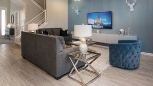 Un salon moderne avec un canapé gris, un pouf bleu, une table basse en verre et un lustre.