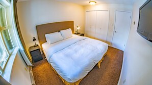Un lit bien fait, avec du linge de lit blanc, une lampe de chevet et une table de nuit. Il y a une fenêtre avec des rideaux, une porte et un téléviseur fixé au mur.
