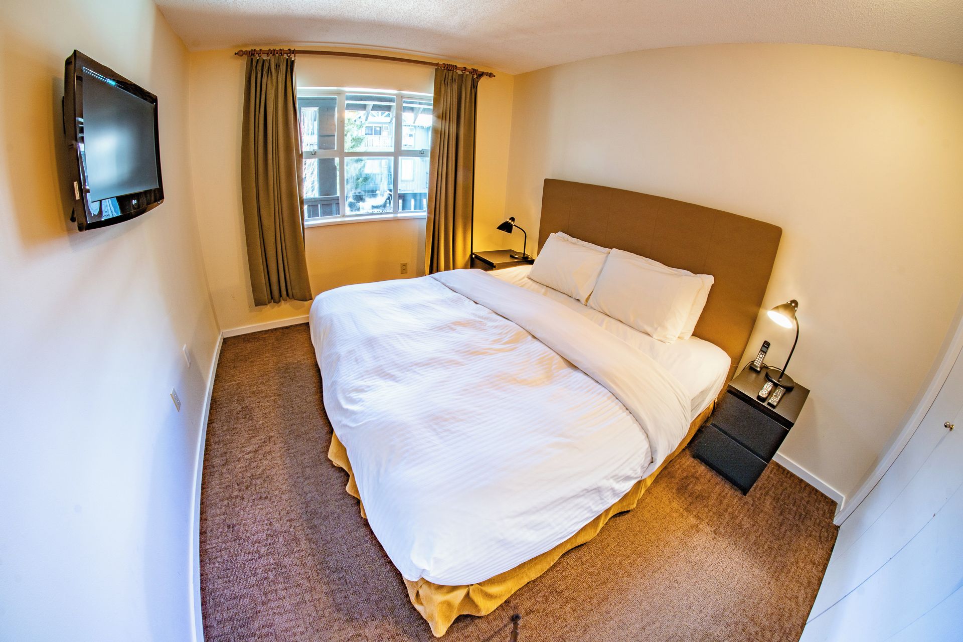 A hotel room with a bed, a TV mounted on the wall, a window with curtains, a bedside lamp, and a nightstand with a phone.