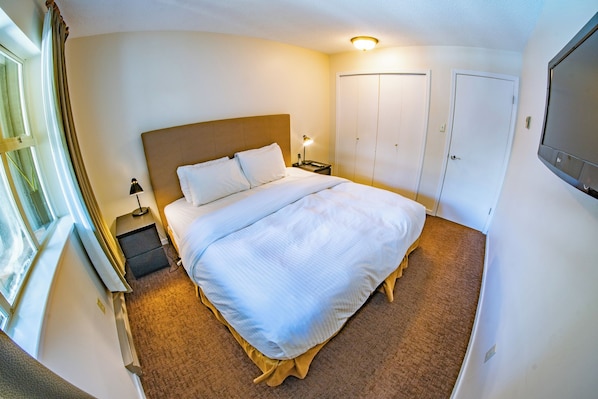 Una cama bien hecha con sábanas blancas, una lámpara de noche y una mesita. Hay una ventana con cortinas, una puerta y un televisor montado en la pared.