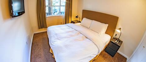 Ein Hotelzimmer mit einem Bett, einem an der Wand montierten Fernseher, einem Fenster mit Vorhängen, einer Nachttischlampe und einem Nachttisch mit Telefon.