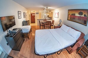 A hotel room with a large bed, a desk with a TV, a chair, a dining table with chairs, and a kitchen area.