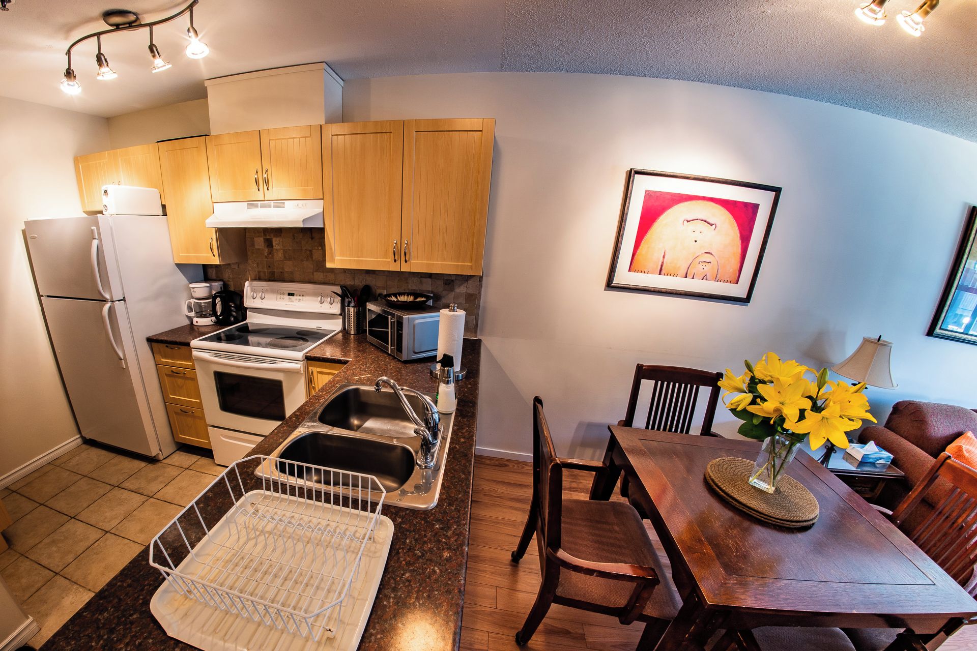 A compact kitchen with a refrigerator, stove, microwave, and sink. There's a dining area with a table and chairs, and a framed picture on the wall.