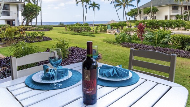 Ein Tisch für zwei, gedeckt mit einem Weinflaschen, blauen Servietten und einem Glas, mit Blick auf einen gepflegten Garten mit Palmen und den Ozean.