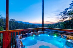 Een hottub met een houten terras en glazen wanden, met uitzicht op een bebost landschap.