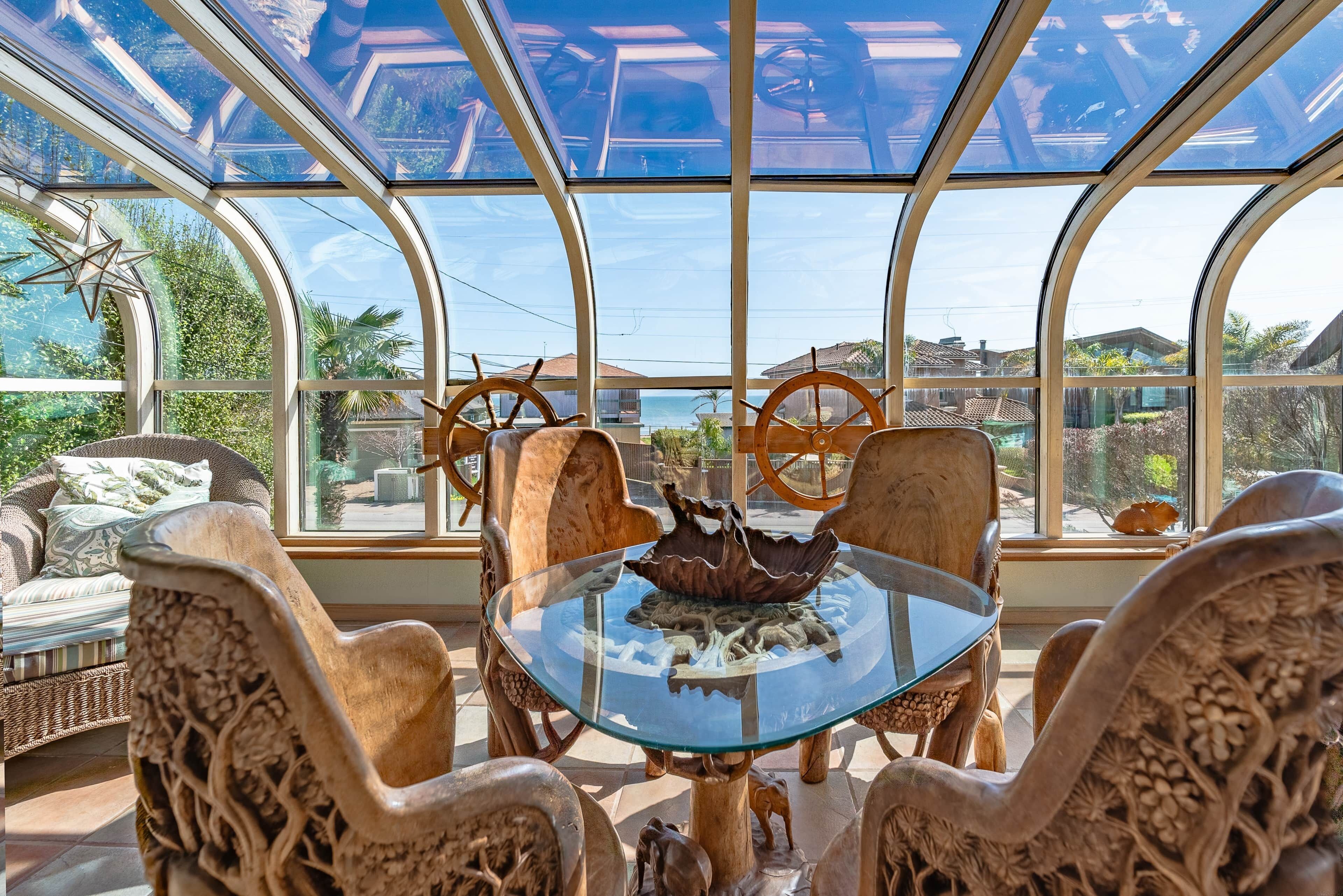 A conservatory with a glass table surrounded by ornate chairs, a decorative wooden ship wheel, and a view of the sea.