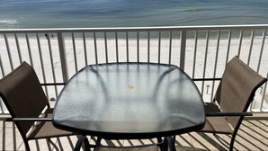 Ein Tisch mit Glasplatte und zwei Stühlen auf einem Balkon mit Blick auf einen Strand.