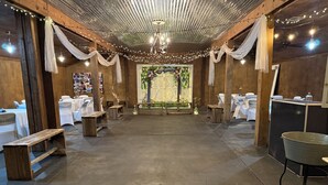 Espaço rústico com paredes de madeira, luzes de corda e um arco floral central. Mesas dispostas com toalhas brancas e cadeiras.