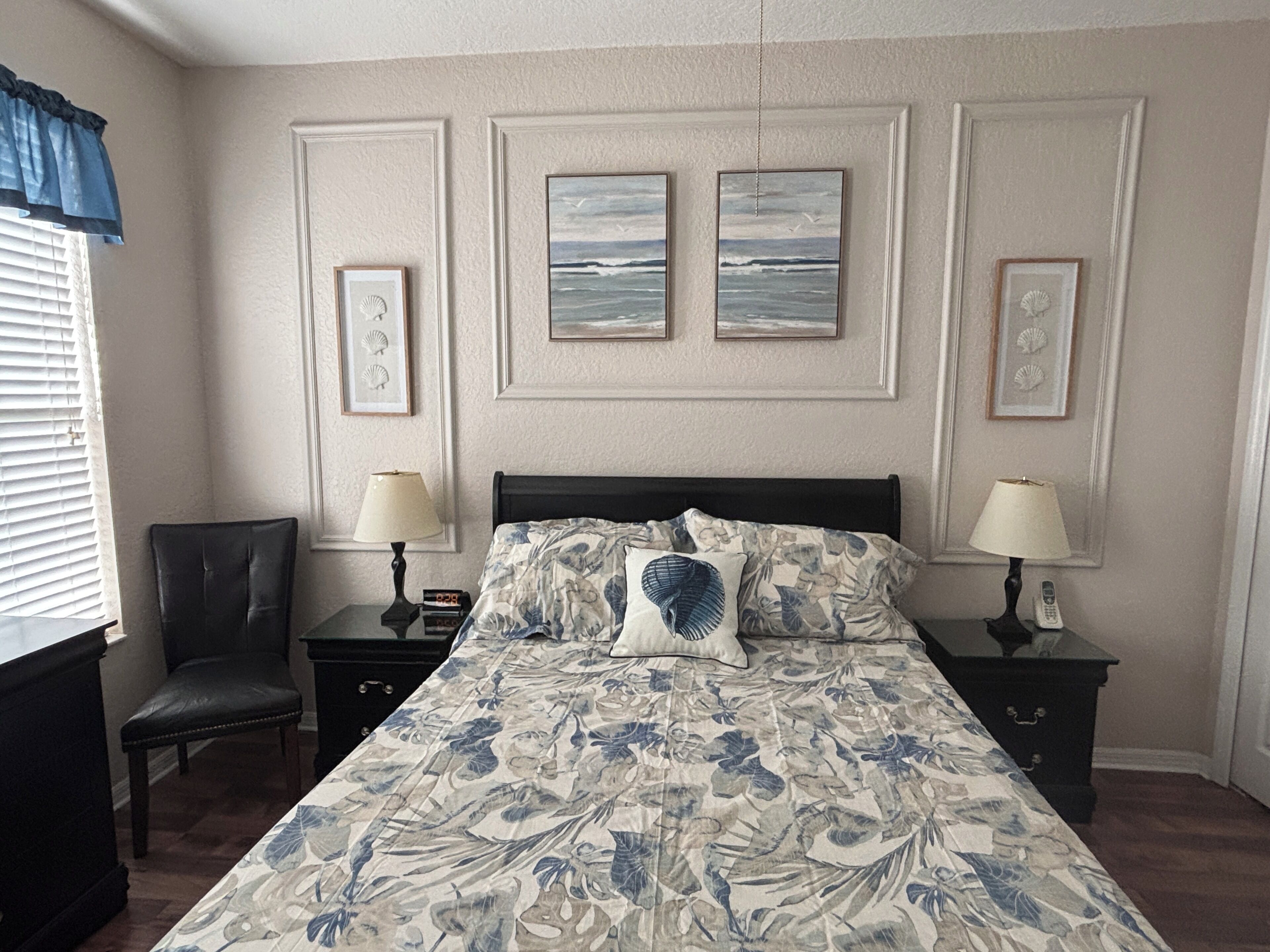 Een slaapkamer met een bed, twee nachtkastjes, een stoel en kunstwerken in een lijst aan de muur.
