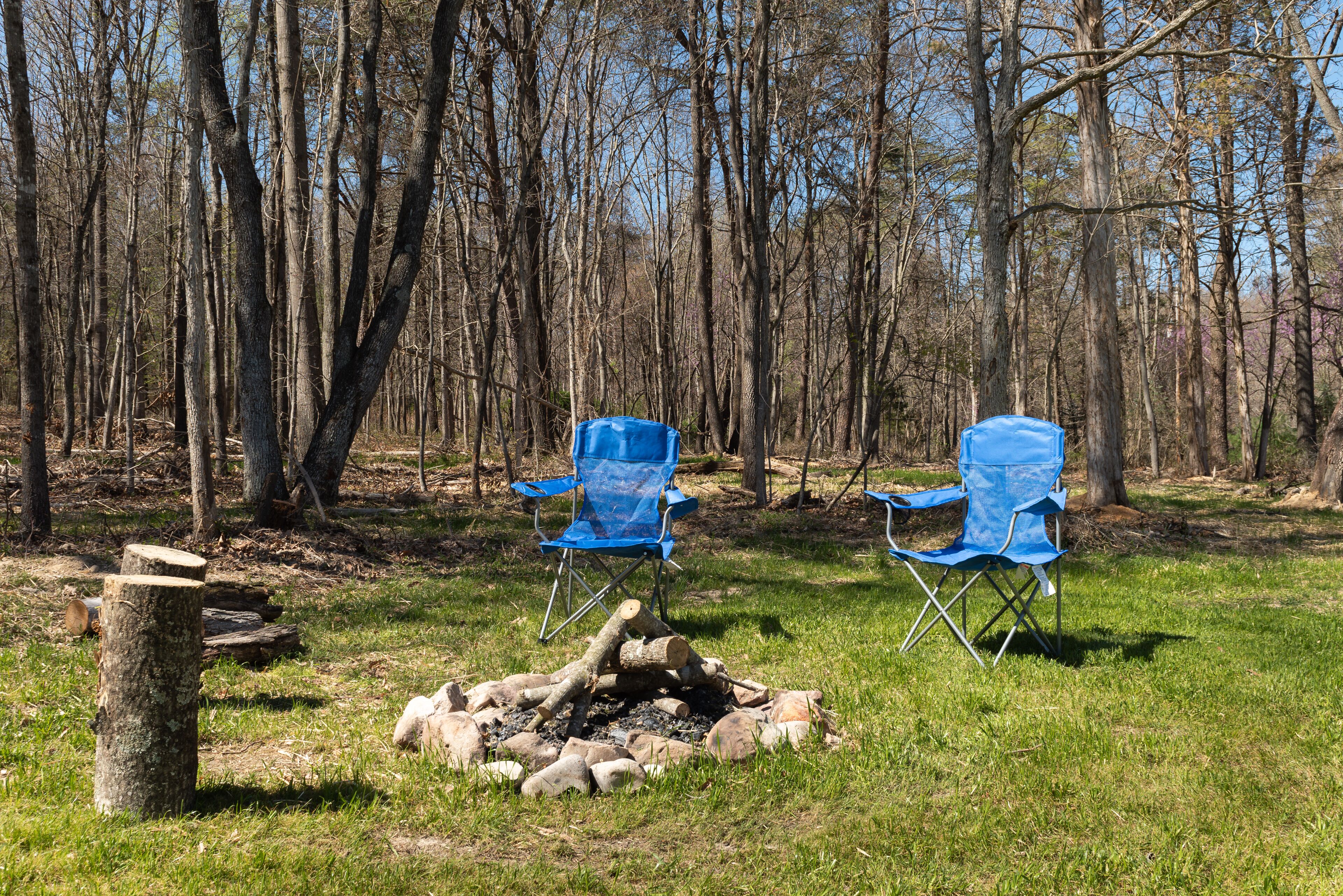 Two blue camping chairs near a campfire in a wooded area.