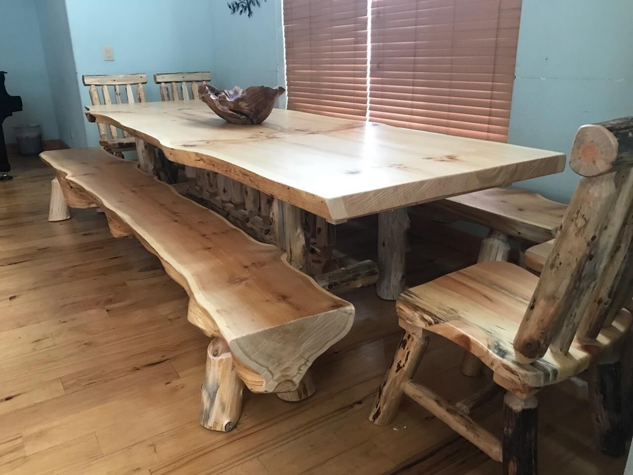 Une table à manger rustique en bois, avec des bancs, un saladier en bois posé sur la table, et une fenêtre avec des stores en arrière-plan.