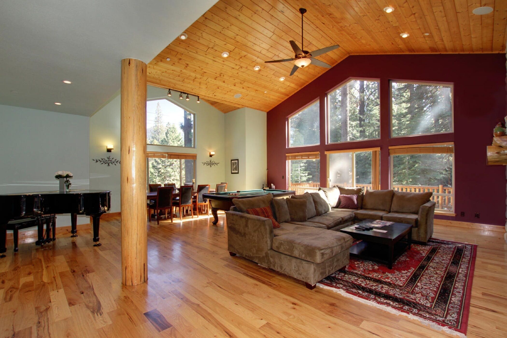 A spacious living room with a wooden ceiling, a pool table, a piano, and a large window overlooking a deck.