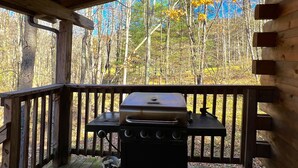 A covered outdoor area with a grill and a table, surrounded by wooden railings and a forest backdrop.