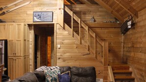 A wooden cabin interior with a staircase, a sofa with cushions, and a sign indicating a ski lodge.