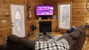 A cozy log cabin interior with a fireplace, a television, a sofa, and a dining table.