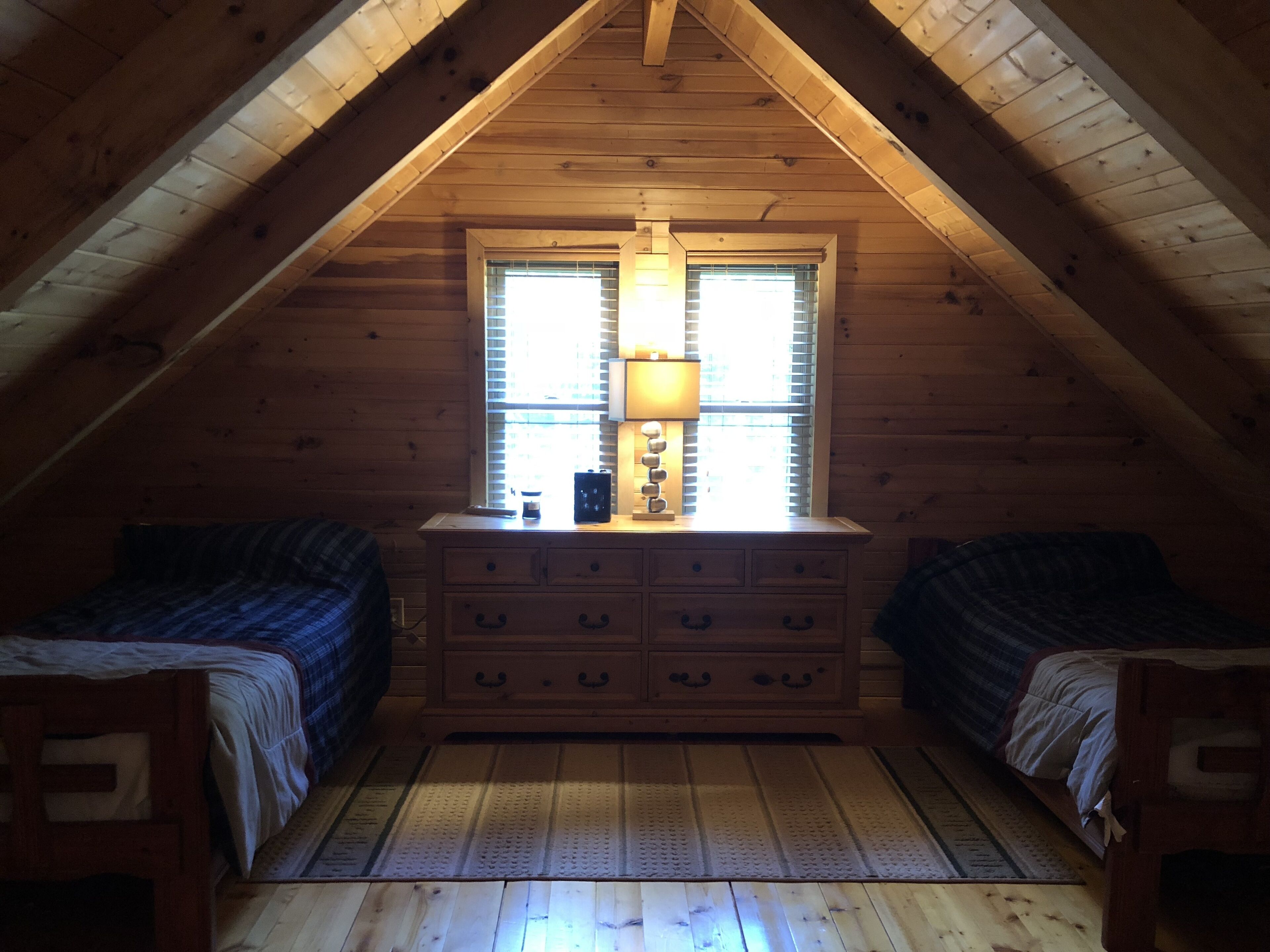 Ein gemütliches Schlafzimmer im Dachgeschoss mit Holzwänden und schrägem Dach. Es gibt zwei Betten mit blauer Bettwäsche, eine Holzkommode mit Schubladen und ein Fenster mit Jalousien.