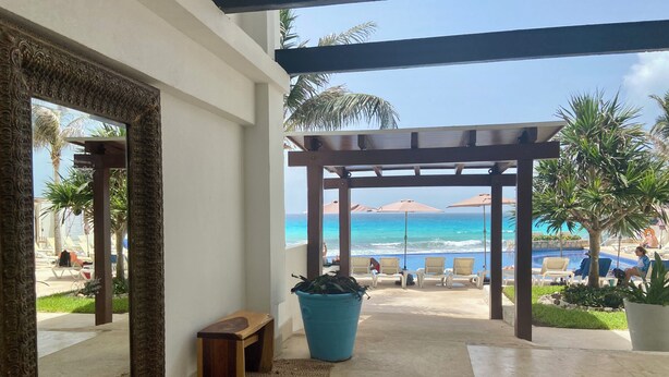 A room with a large mirror, a wooden bench, a blue pot, and a view of a beach with lounge chairs and umbrellas.