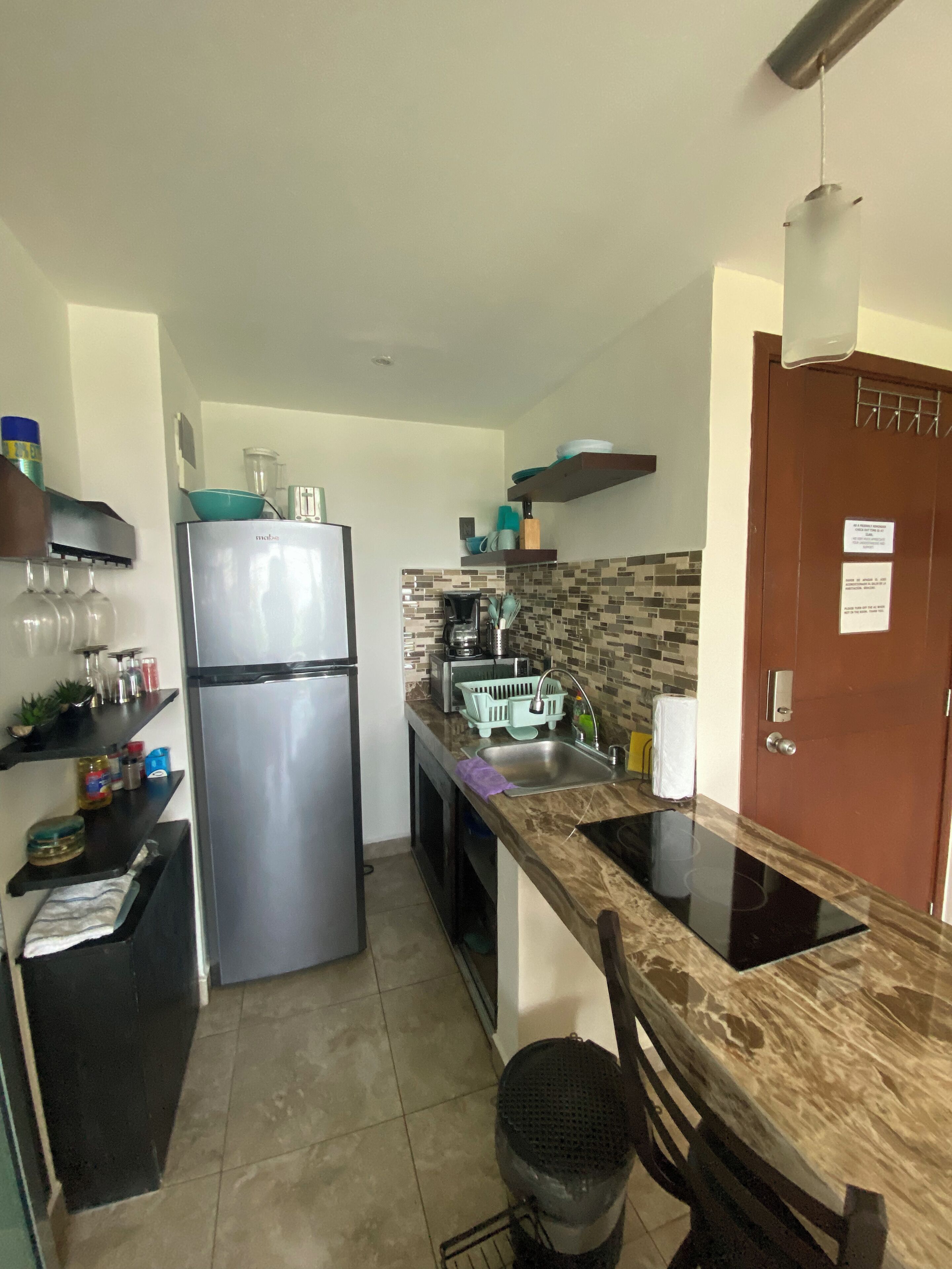 Cocina compacta con refrigerador de acero inoxidable, horno empotrado y pileta. Hay estantes con utensilios de cocina y un ventanal sobre la pileta.