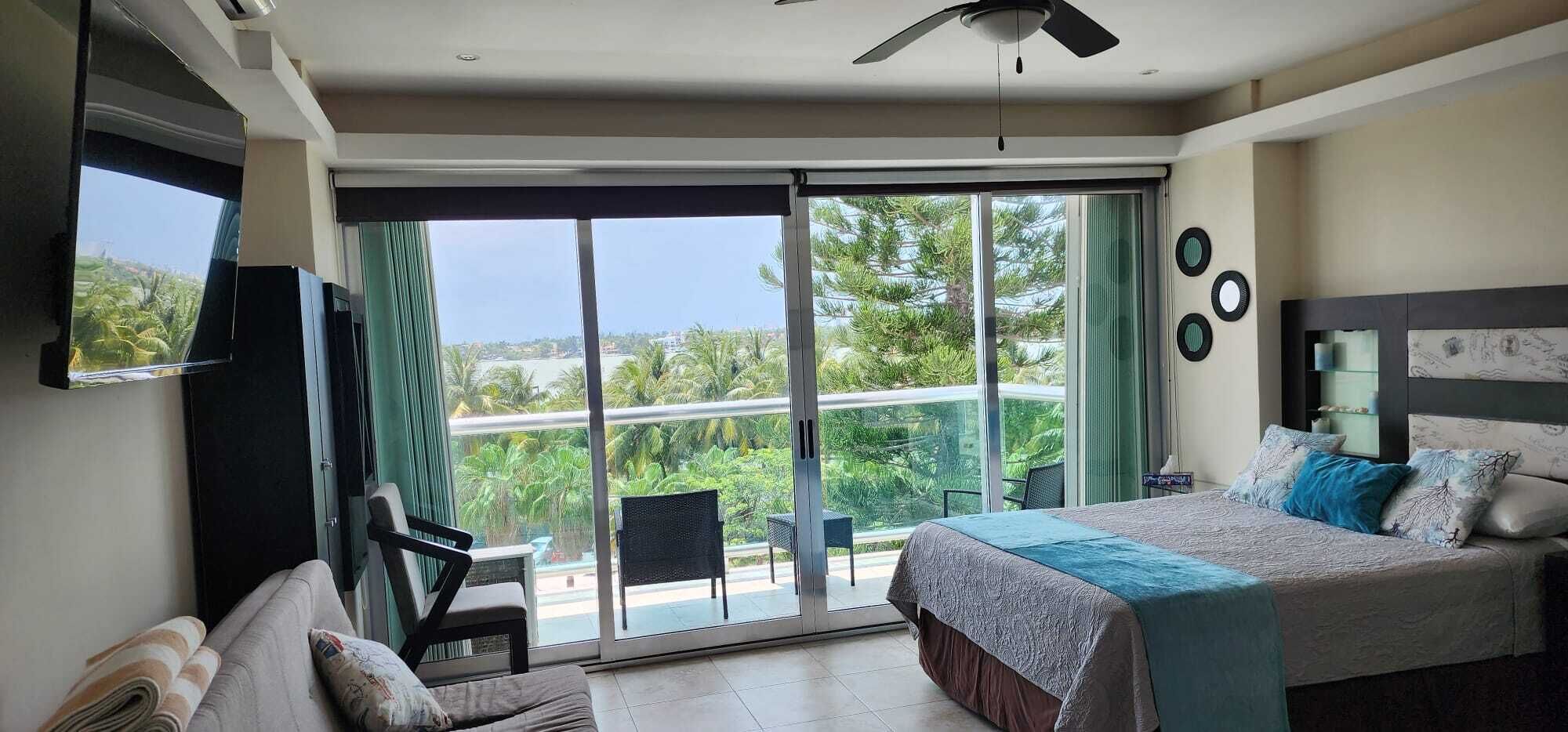 Une chambre avec un lit, une télévision, un ventilateur de plafond et un balcon offrant une vue sur la verdure.