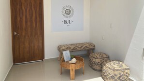 A small, clean room with a wooden door, a wall-mounted sign reading "KU SPA", a bench, and two patterned ottomans.