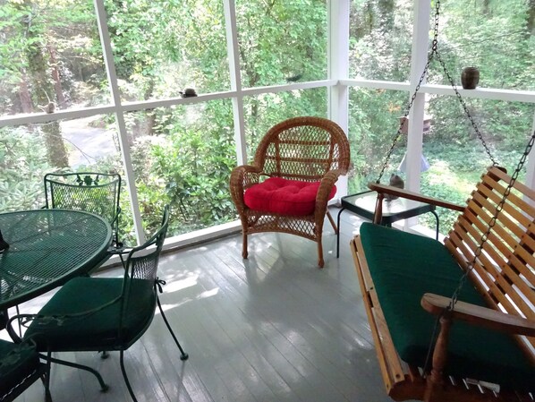 Relax in a peaceful space with a wooden swing chair, wicker chair, and metal table amidst lush greenery views.