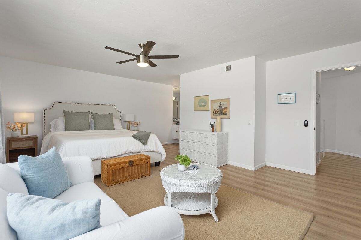 Een slaapkamer met een bed, een witte bank, een rieten bijzettafeltje, een plafondventilator en een dressoir.