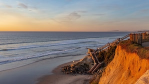 Blick auf die Küste mit einer Holztreppe, die zum Strand führt, einer felsigen Uferlinie und einem klaren Himmel.