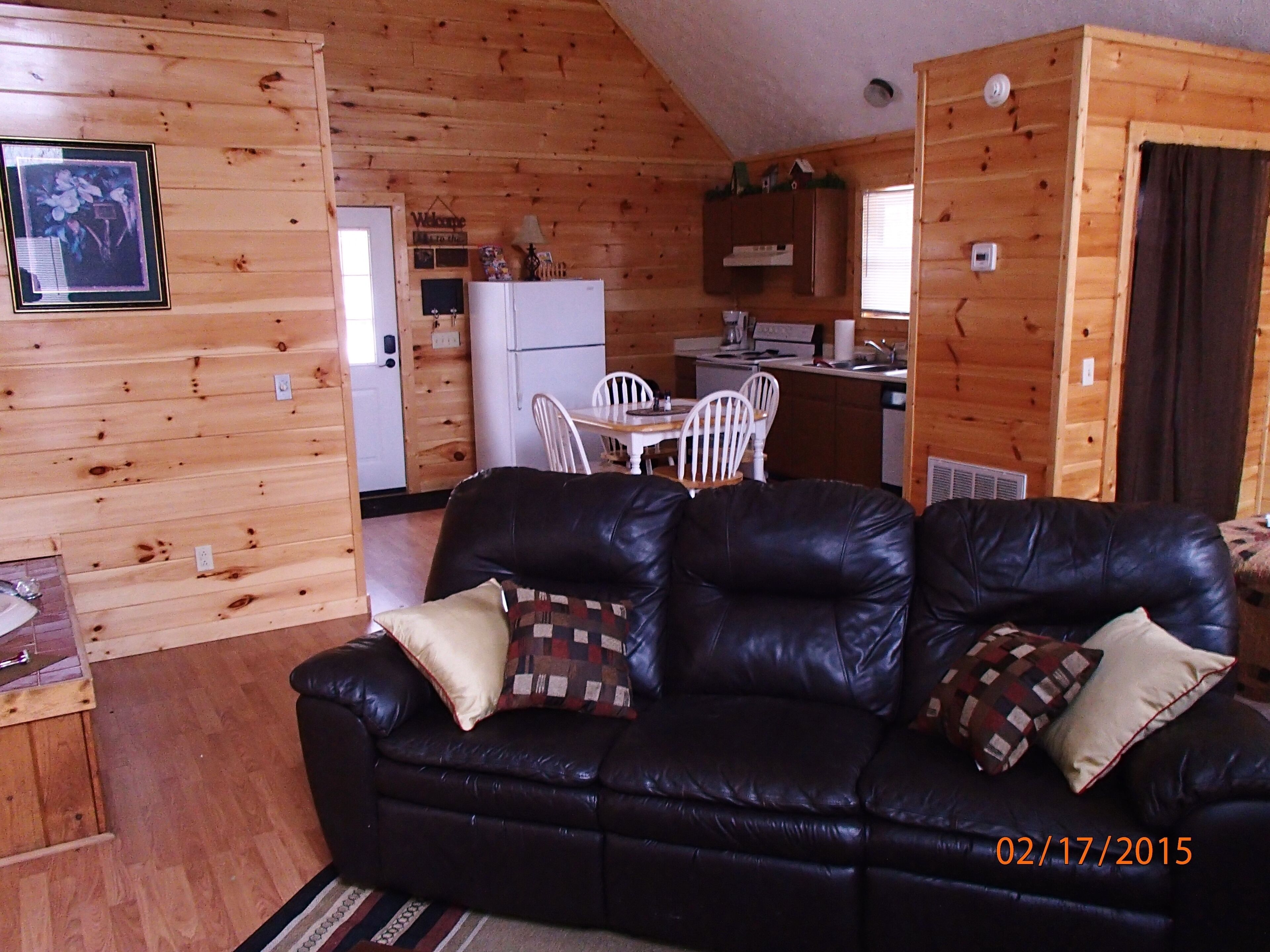A cozy cabin interior with a black leather sofa, wooden walls, a dining area with white chairs, and a kitchen with a refrigerator and stove.