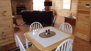L’intérieur d’un chalet accueillant, avec une cheminée en pierre, un canapé noir, une table à manger avec des chaises et un coin cuisine équipé d’un évier et d’une fenêtre.
