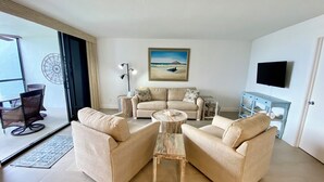 A living room with a beige sofa set, a coffee table, a side table, and a painting of a beach scene. - Water View of Marco Pass, Heated Pool & Marina-Newly Updated 5th Floor 1Br/1Ba (Marco Island)