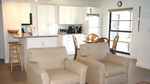 A modern kitchen with white cabinets, a built-in oven, and a dining area with two beige armchairs and a wooden dining table. - Water View of Marco Pass, Heated Pool & Marina-Newly Updated 5th Floor 1Br/1Ba (Marco Island)