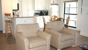 Una cocina moderna con armarios blancos, horno empotrado y un comedor con dos sillones color beige y una mesa de madera.