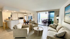 A modern living room with a sofa, chairs, and a coffee table. There is a kitchen area with white cabinets and a dining table. The room has large windows with a view of the outdoors. - Water View of Marco Pass, Heated Pool & Marina-Newly Updated 5th Floor 1Br/1Ba (Marco Island)