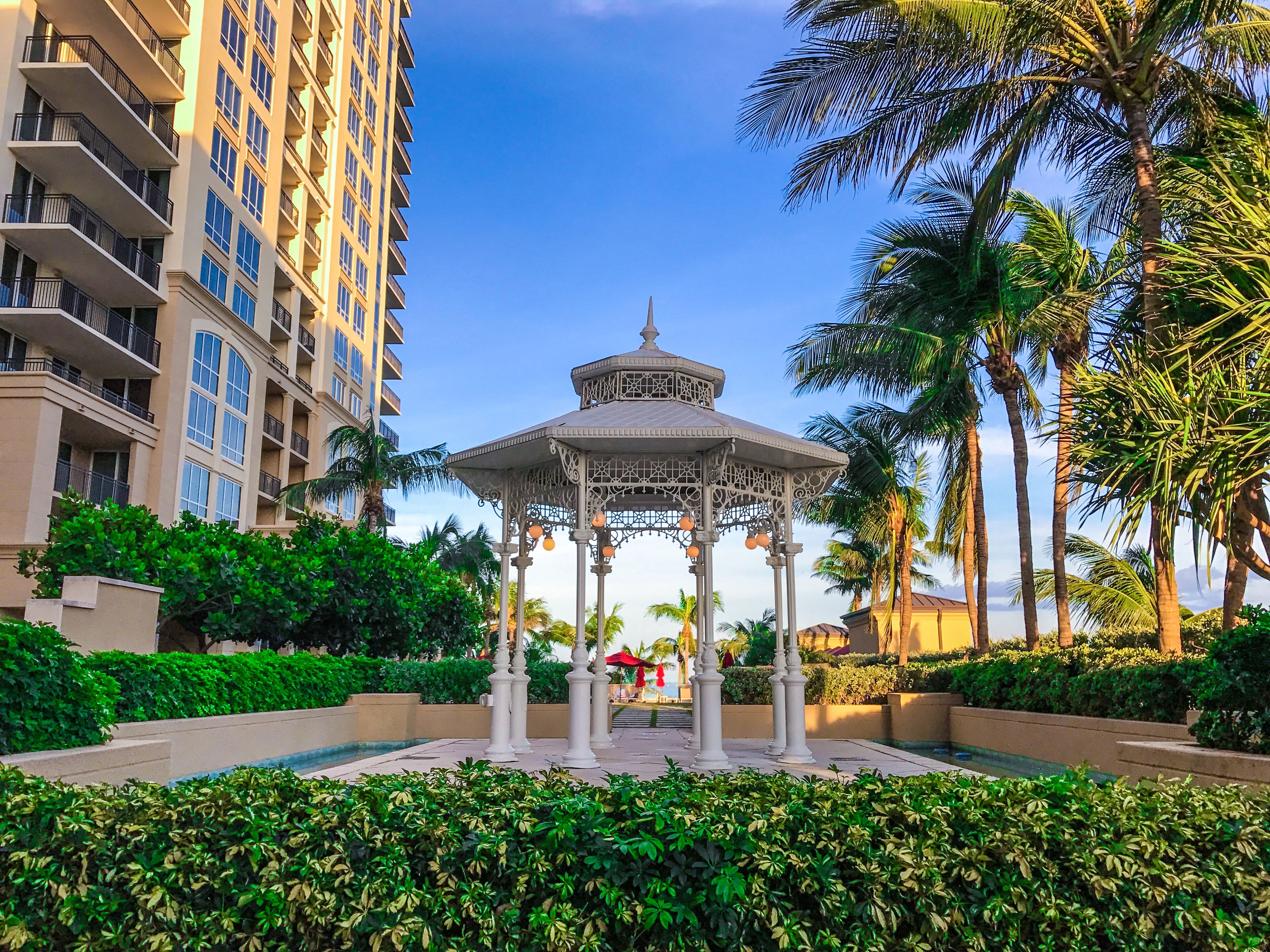 A gazebo surrounded by lush greenery and palm trees, with a high-rise building in the background.