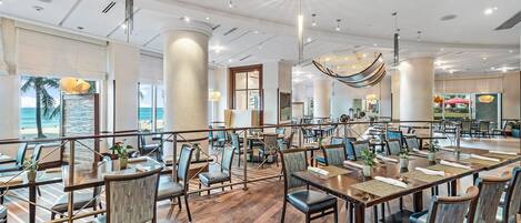 A spacious dining area with multiple tables and chairs, large windows with a view of the beach, and a modern ceiling design.