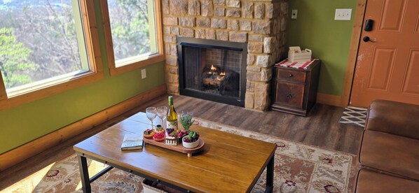 Una habitación acogedora con chimenea de piedra, una mesa de madera con una bandeja de postres y vistas a través de un ventanal grande.