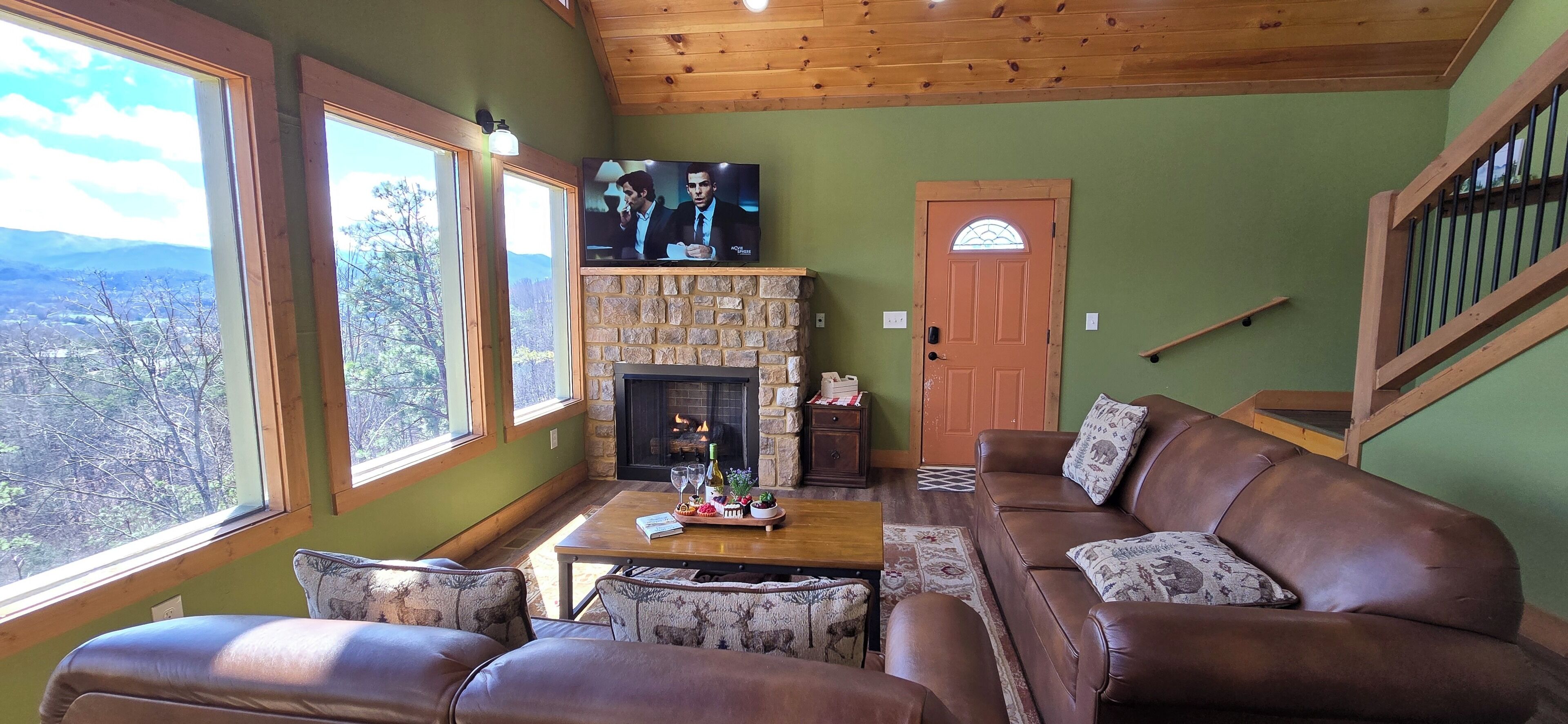 A cozy living room with a stone fireplace, leather sofa, and large windows offering a view of the outdoors.