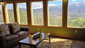 Una habitación con un sofá de cuero marrón, una mesa de centro de madera con una botella y vasos, y vistas a montañas a través de amplios ventanales.