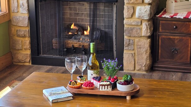Una mesa de madera con una bandeja de postres, una botella de vino y dos copas. Hay un libro sobre la mesa, y una chimenea al fondo.