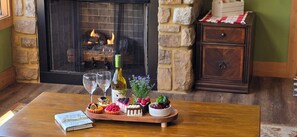 Ein Holztisch mit einer Auswahl an Desserts, einer Weinflasche und zwei Gläsern. Auf dem Tisch liegt ein Buch, im Hintergrund ist ein Kamin zu sehen.