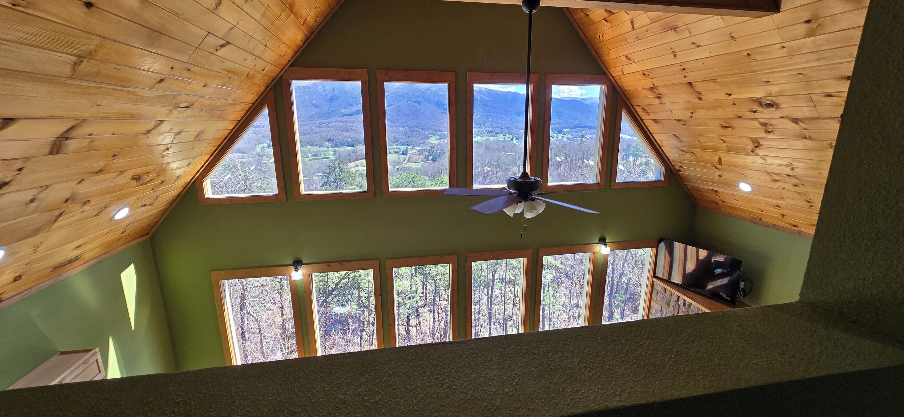 Una habitación con techo inclinado, varias ventanas, ventilador de techo y vistas a un paisaje boscoso.