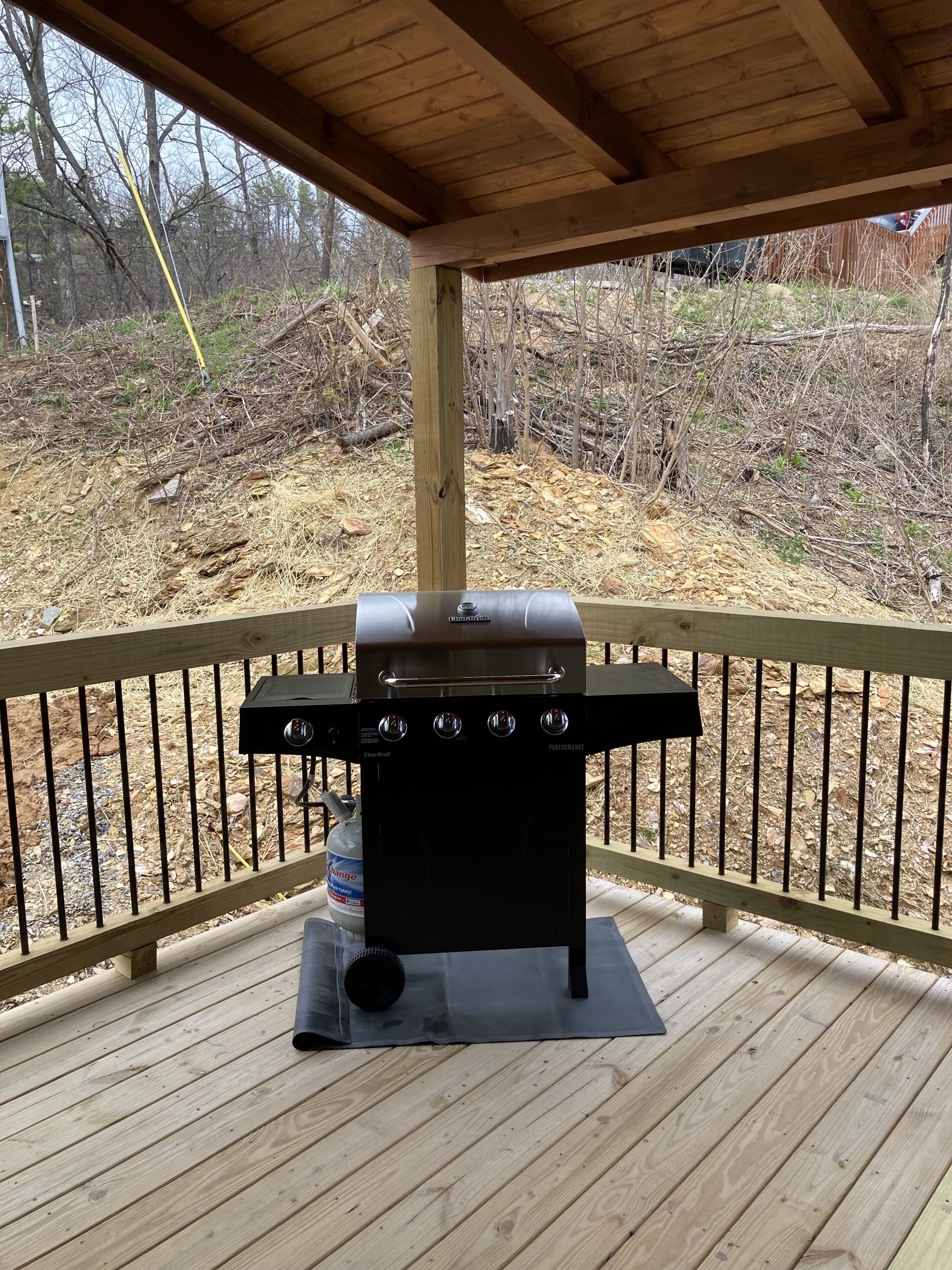 Un barbecue installé sur une terrasse en bois dotée d’une rambarde, le tout entouré d’une zone boisée.
