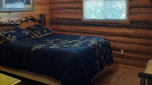 Une pièce de style cabane en bois, avec un lit, deux fenêtres, un ventilateur de plafond et un tableau encadré au mur.
