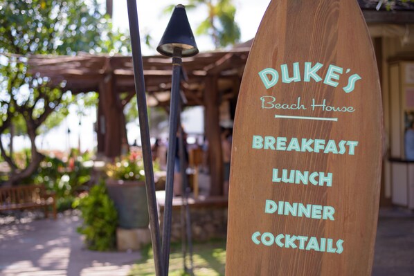 Ein hölzernes Schild mit dem Schriftzug 'DUKE'S Beach House' und der Aufzählung der angebotenen Leistungen: 'FRÜHSTÜCK, MITTAGESSEN, ABENDESSEN, COCKTAILS'.