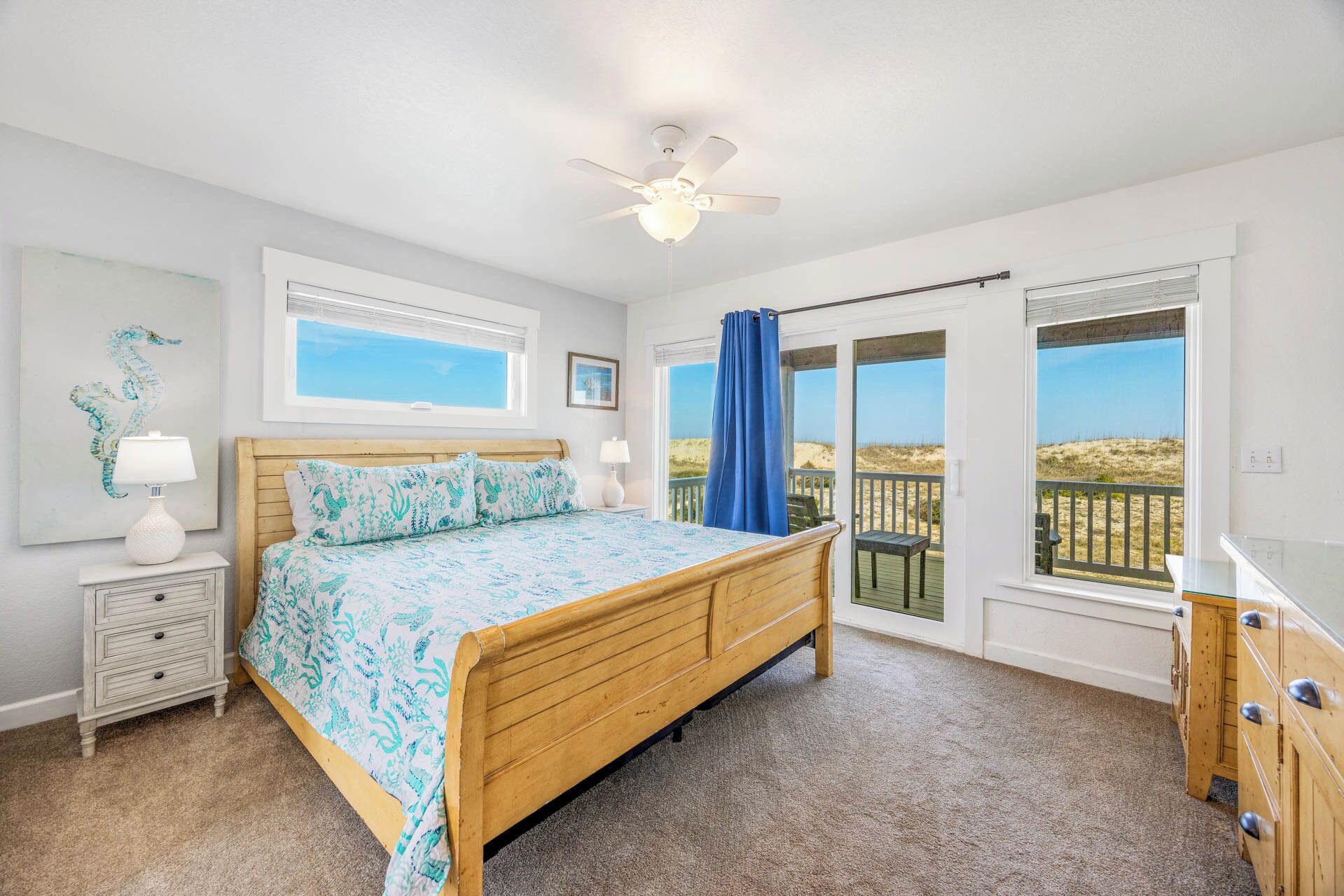 Une chambre avec un lit en bois, un ventilateur de plafond, une table de chevet avec une lampe, une commode et de grandes fenêtres donnant sur la plage.
