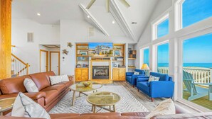 A spacious living room with a fireplace, a large window overlooking the ocean, and a cozy seating area. - Premier Oceanfront Home in Waves w/ Private Pool, Hot Tub, Game Room, Tiki Bar (Waves)
