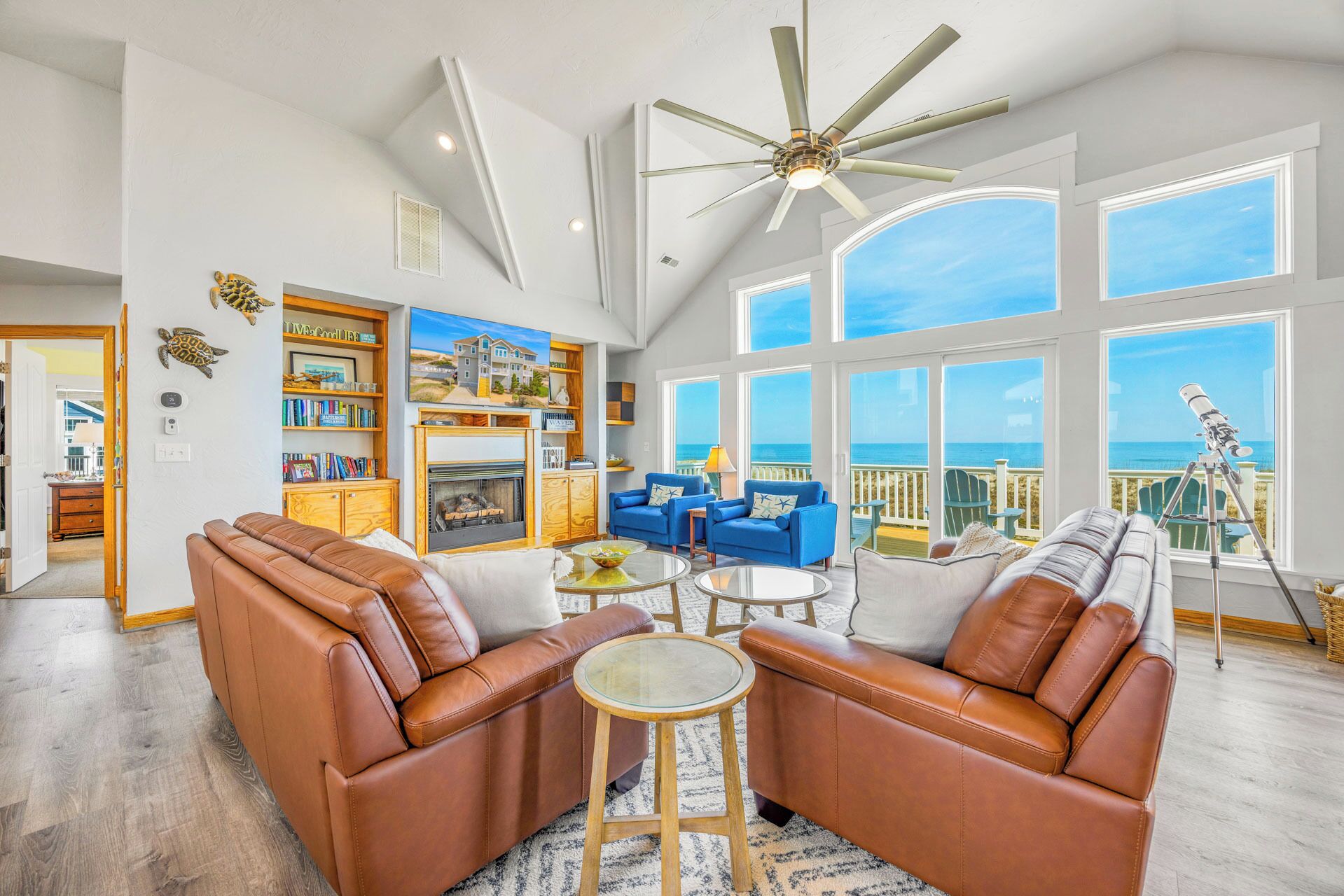 A spacious living room with leather sofas, a fireplace, a large window with a view of the ocean, and a ceiling fan.